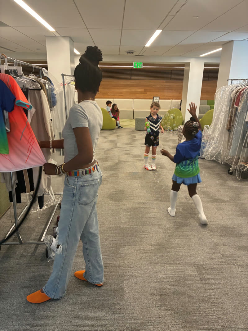 A young stylist organizes garments on a wardrobe rack while children play nearby