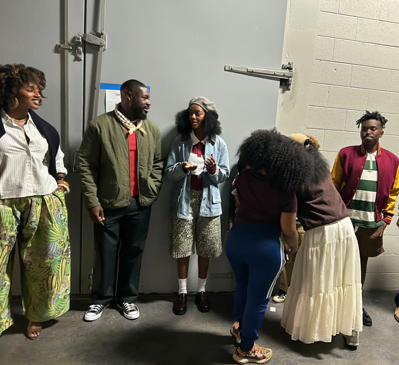 Participants and mentors gather backstage while a stylist checks each look