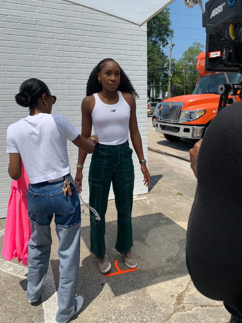 A stylist fits a garment during an outdoor photo shoot alongside a production camera